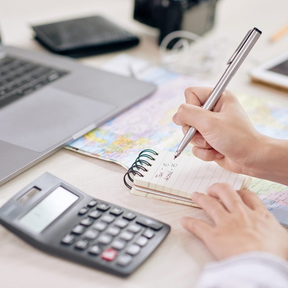 Crop shot of woman taking notes in notepad sitting at desk with laptop and calculating machine
