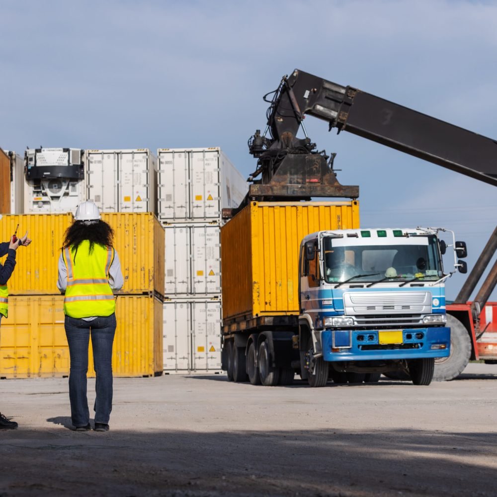 Two female workers in safety vests and hard hats oversee container loading operations at a shipping yard. A crane lifts a yellow container onto a truck, highlighting teamwork and global logistics.